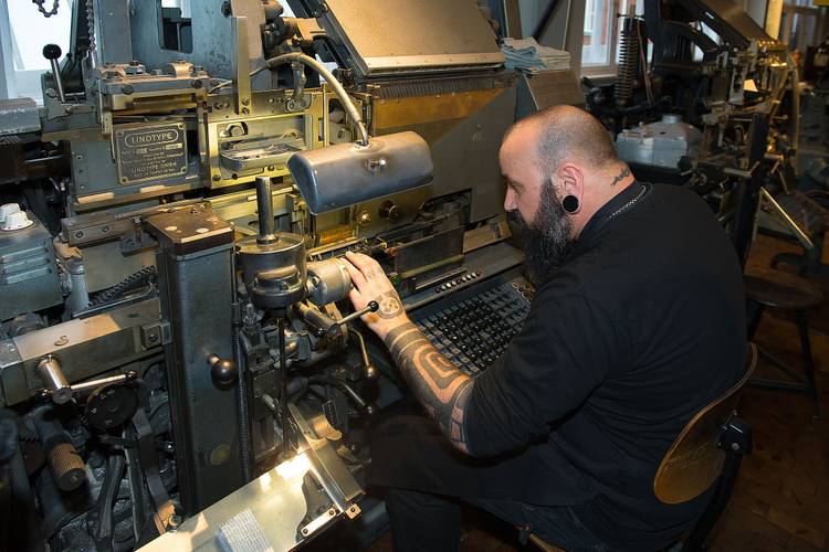 Vorführungen an der Linotype veranschaulichen das automatisierte Setzen. Die Linotype wird auch der „Eiserne Kollege“ genannt. – Ein Mann arbeitet an einer großen, schwarzen Setzmaschine.