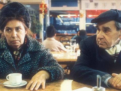  – Zwei Personen sitzen an einem Tisch in einem Cafe. Links eine weiblich gelesene Person mit Fellhut, rechts ein maennlich gelesener Mann mit Schiebermuetze.