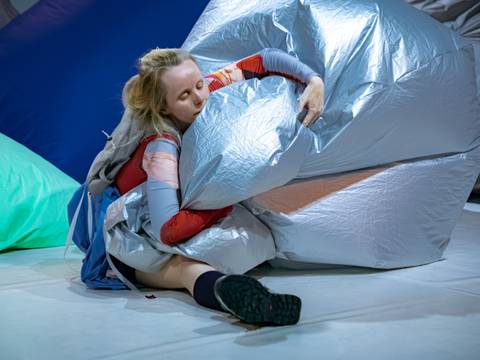 in the air – Person in bunter Kleidung umarmt großes silbernes Kissen, sitzt auf Boden mit buntem Hintergrund aus blauen und grünen Kissen.