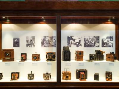 Erste Handkameras: Die Ausstellung „Faszination des Augenblicks“ zeigt die Entwicklung der Fotografie. – Eine große Vitrine aus dunklem Holz zeigt verschiedene historische Kameras mit Holzgehäuse. Im Hintergrund hängen einige Schwarzweißaufnahmen.