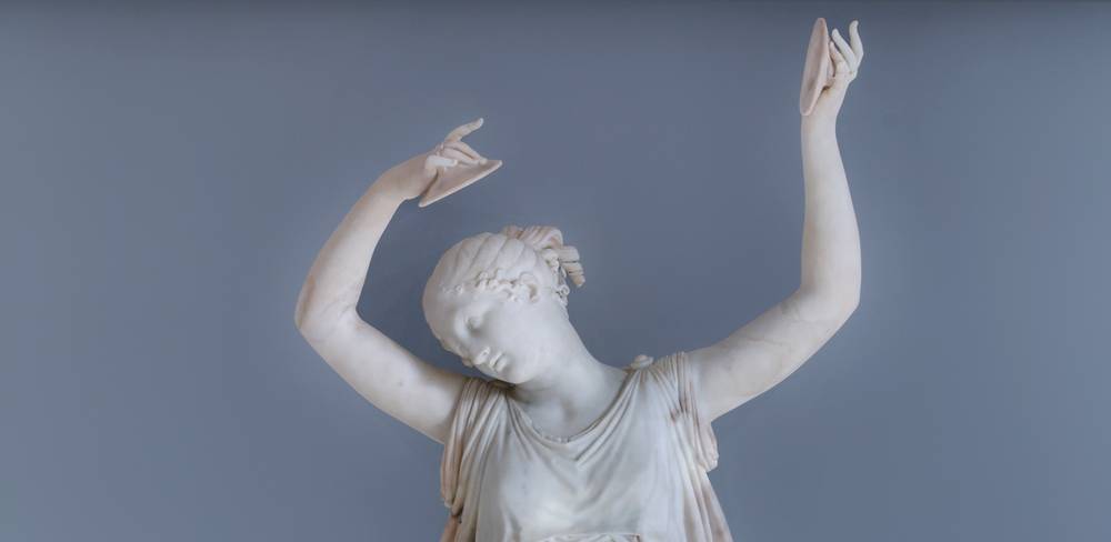 Antonio Canova, Tänzerin mit Zimbeln – Marmorstatue einer Person mit erhobenen Armen, Kopf geneigt. Fließende Gewandung, geschlossene Augen. Hintergrund graublau.