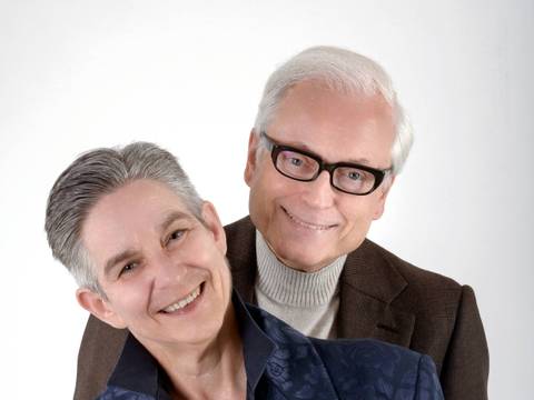 Sigrid Grajek und Hans-Jürgen Schatz – Zwei lächelnde Personen; die vordere trägt ein florales Sakko, die hintere eine Brille und einen Pullover unter einem Sakko.