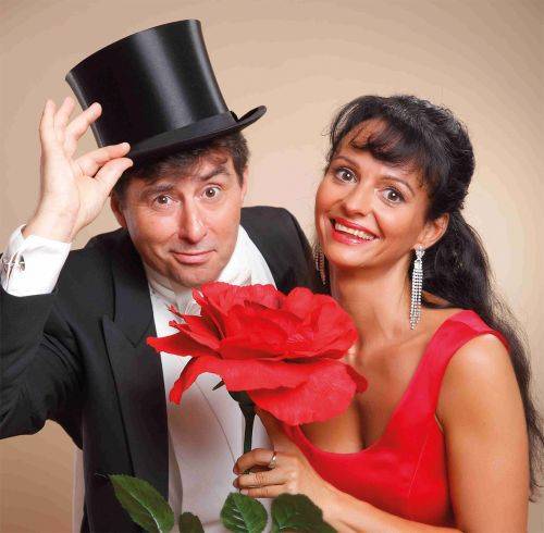Eine lächelnde Frau mit roten Kleid und Ohrringen, ein Mann hält eine große rote Rose und hebt einen Zylinderhut.