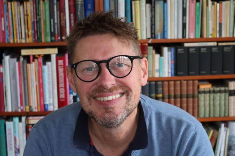 Person mit Brille lächelt vor einem Bücherregal mit bunten und schwarzen Büchern.