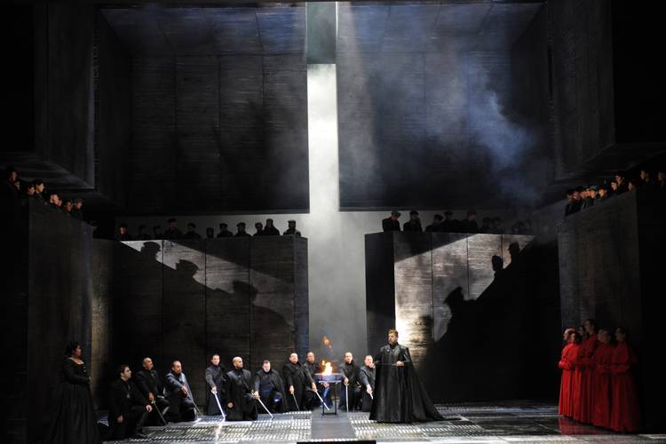 Don Carlo – Dunkle Bühne mit vielen Personen, eine Person in schwarz steht vorne, mehrere Personen in schwarzen und roten Gewändern im Hintergrund, Lichtstrahl von oben.