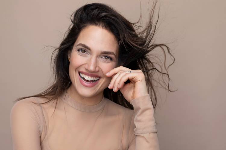 Marina Maximilian – Lachende, weiblich gelesene Person mit langen, wehenden Haaren und transparentem Oberteil vor neutralem Hintergrund.