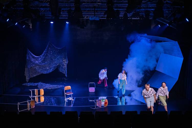 "Die Fische" am Theater Strahl – Dunkle Bühne mit drei Personen und einer im Nebel gehüllten Konstruktion. Drei umgefallene Stühle, Netzstruktur auf der linken Seite.