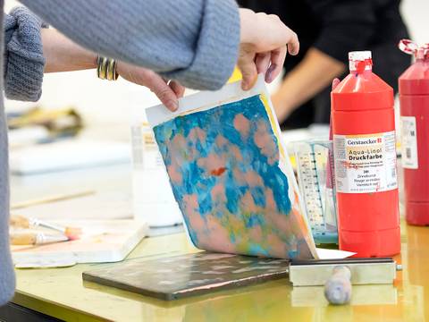  – Person hält ein Blatt mit buntem Druck. Zwei rote Flaschen mit Aufschrift "Gerstaecker Aqua-Linol Druckfarbe". Malutensilien auf gelbem Tisch.