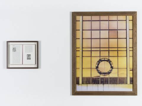 Sophie Calle, DDR Wappen (Palast der Republik), 1996, Farbfotografie und Buch, 2-teilig, 120 x 90 x 4 cm (Bild), Hamburger Bahnhof – Nationalgalerie der Gegenwart, Dauerleihgabe der Commerzbank – Ein kleines gerahmtes Dokument links und ein großes Foto eines Fensters mit gelbem Licht und kreisförmigem Metallmuster rechts, beide an einer weißen Wand.