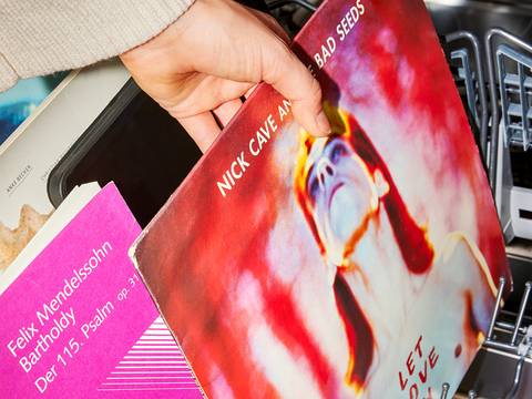  – Hand hält Schallplatte mit "NICK CAVE AND THE BAD SEEDS", daneben Buch mit "Felix Mendelssohn Bartholdy Der 115. Psalm op. 31" in Spülmaschine.
