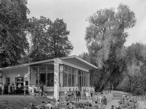 Milchhäuschen am Weißensee von Ludmilla Herzenstein, um 1978, Berlin-Weißensee, Parkstraße 33a – Schwarz-Weiß-Fotografie des Milchhäuschens in Berlin-Weißensee, vor dem viele Menschen in kleinen Grüppchen an Tischen im Außenbereich sitzen. Das Milchhäuschen wird von hohen Bäumen gesäumt.