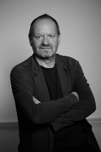 Person mit Bart, gekreuzte Arme, dunkles Oberteil und Blazer vor neutralem Hintergrund. Schwarzweiß-Fotografie.