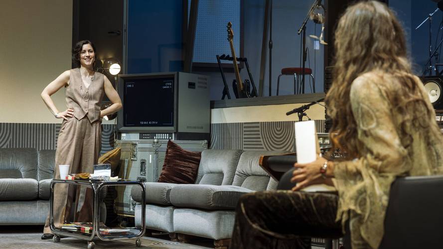 Stereophonic – Zwei Personen in einem Raum mit Sofas, Musikinstrumenten und einem Bildschirm mit der Aufschrift "C100". Eine Person sitzt, die andere steht lächelnd.