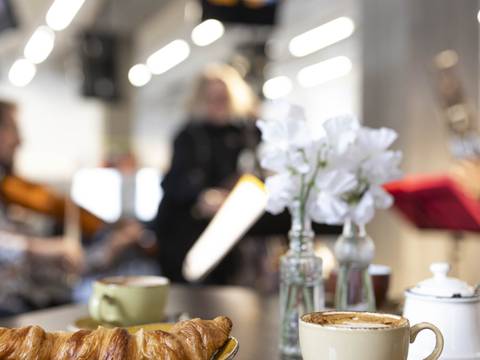  – Croissant und Kaffee auf einem Tisch, im Hintergrund spielen Musiker