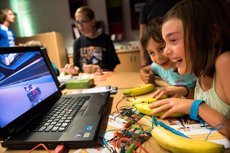 Mit leitfähigen Materialien, wie Bananen, Computer und Videospiele steuern. – Kinder sitzen an einem Tisch vor einem Laptop. Auf dem Bildschirm ist ein Spiel mit einem Rennauto zu sehen. Die Kinder steuern das Spiel mit Bananen, die auf dem Tisch liegen und über Kabel und ein Gerät mit dem Laptop verbunden sind.