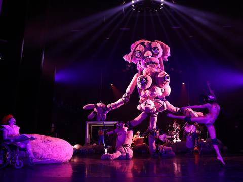  – Bühnenperformance mit großer, farbenfroher Kreatur in der Mitte, umgeben von Performer*innen in futuristischen Kostümen, bühnenhafte Beleuchtung.