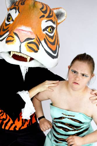 Person im Tigerkostüm mit großem Kopf hält die Schulter einer Frau. Die Frau trägt ein schulterfreies Kleid in Türkis mit schwarzem Muster.