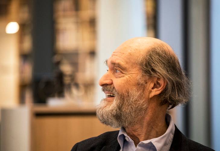 Arvo Pärt – Ältere Person mit Bart und Halbglatze, blickt lächelnd nach links. Hintergrund unscharf mit Regalen.