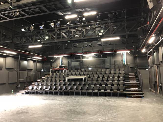 Leerer Theaterraum mit schwarzen Stuhlreihen und Podesten; helle Bühnenbeleuchtung an der Decke.