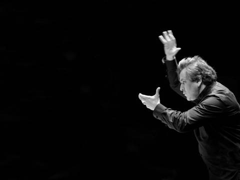 Sir Antonio Pappano, Chefdirigent des London Symphony Orchestra – Am rechten Bildrand ist vor schwarzem Hintergrund ein Mann zu sehen, der beim Dirigieren die Arme vor sich in die Höhe hält.