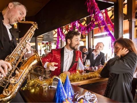  – Person in rotem Paillettenjackett lacht an einer Bar, spielt Saxophon; weitere Personen feiern mit Partyhueten und Dekorationen, Geraeuschempfindlichkeit