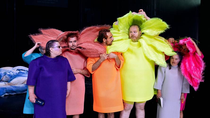 Sechs Personen in bunten Kostuemen mit großen, blütenartigen Halskrausen. Farben sind lila, rosa, orange, neongelb, hellgrau und pink. Schwarz als Hintergrund.