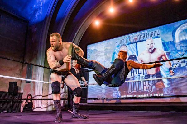 Odeum Pro Wrestling – Zwei Personen ringen im Ring, eine Person in schwarzer Hose hebt eine andere Person. Hintergrund: Projektion mit Text "Kluth vs Oberhau".