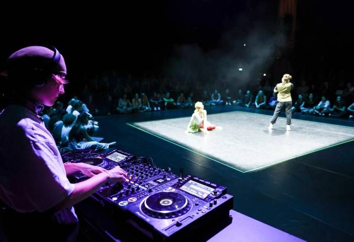 Tanzbegegnungen bei der ARENA – Ein DJ legt an einem Mischpult auf. Im Hintergrund ist ein hell beleuchteter Cypher zu sehen, in dem sich zwei Personen befinden. Das Publikum sitzt rundherum.