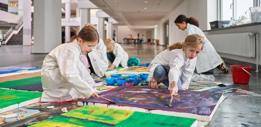 Familienworkshop in der Neuen Nationalgalerie – Kinder in weißen Kitteln malen mit Pinseln auf großen Leinwänden auf dem Boden in einer hellen Halle. Farbtöpfe sind um sie herum verteilt.