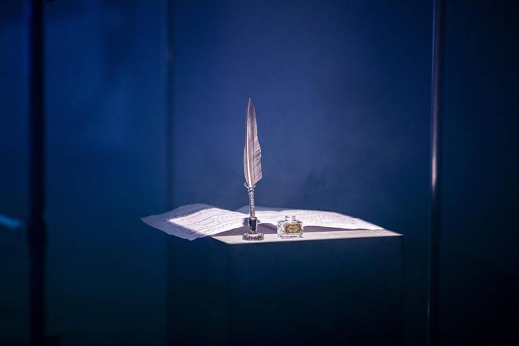 Feder in Halter, Tintenfass und Papier auf Tisch in dunkler Umgebung, bläuliches Licht.