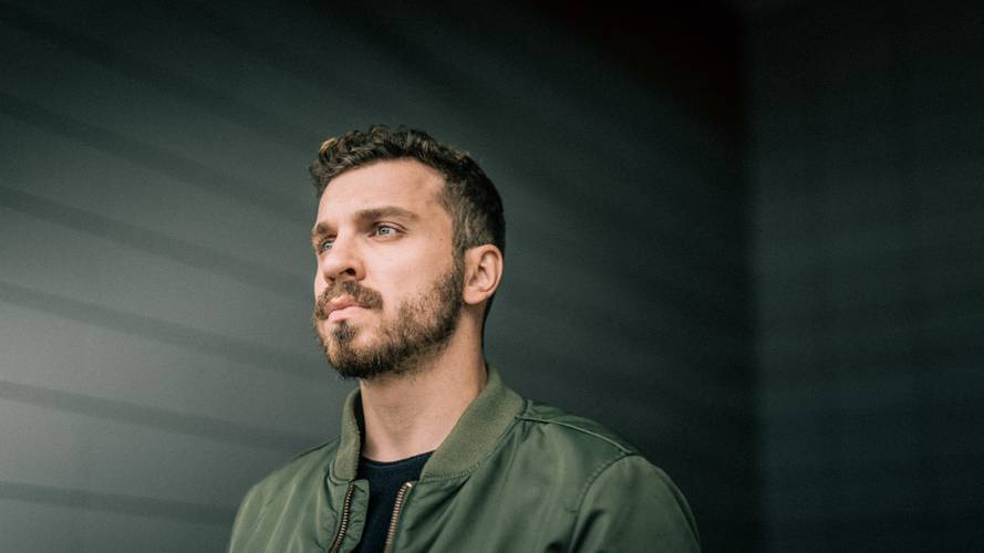 Edin Hasanovic – Person mit Bart, schaut nach links, trägt grüne Jacke, dunkles T-Shirt. Hintergrund mit horizontalen Linien, weich beleuchtet.