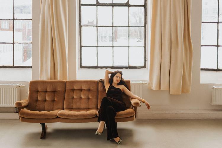 Anna Margolina – Anna Margolina sitzt in einem lofartigen Raum mit großen Fenstern und cremefarbenen Vorhängen auf einem braunen Vintage-Velour-Sofa. Sie trägt ein schwarzes Kleid und Pumps.