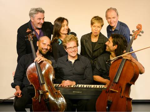  – Sieben Personen mit Instrumenten; zwei Cellist*innen im Vordergrund, eine Person am Roland-Klavier, die restlichen im Hintergrund stehend, lächelnd.