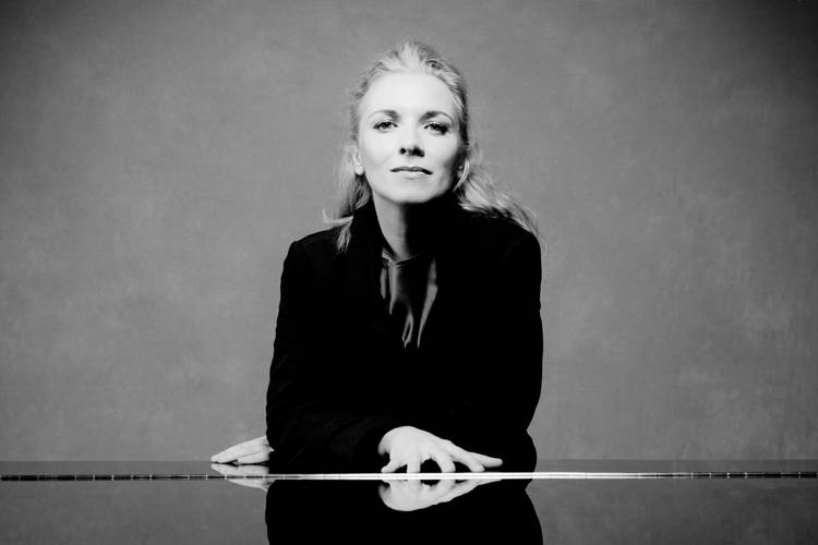 Die Pianistin Tamara Stefanovich – Eine Frau mit blonden Haaren in einem schwarzen Samtblazer steht hinter einem zugeklappten Klavierflügel und schaut in die Kamera.