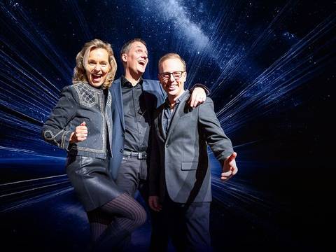  – Drei gut gelaunte Personen in eleganter Kleidung vor einem Hintergrund mit Weltraum-Motiv. Alle lachen und machen Daumen-hoch-Gestik.