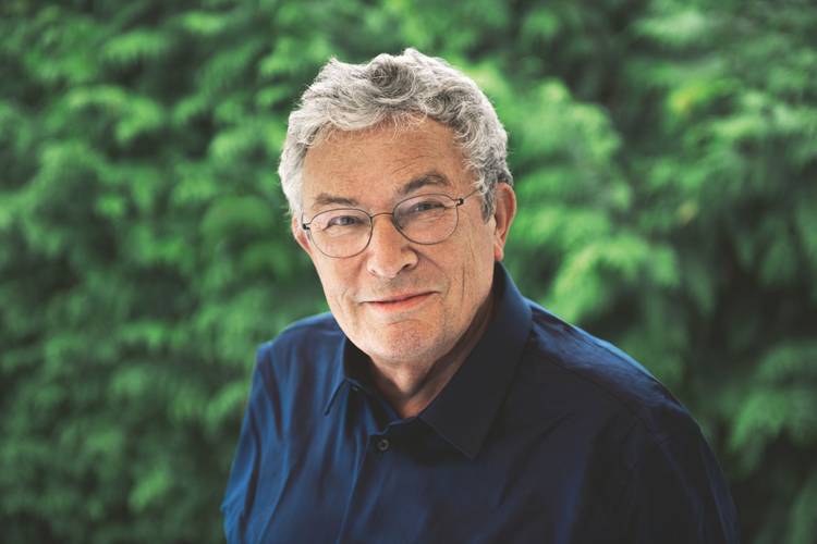 Charles Lewinsky – Ältere Person mit Brille und blauem Hemd vor grünem, unscharfem Hintergrund mit Blättern.