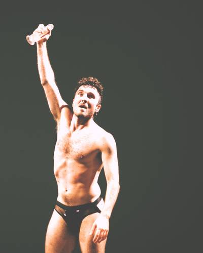 Performer mit nacktem Oberkörper auf dunkler Bühne, Arm erhoben, transparenter Dildo in der Hand.