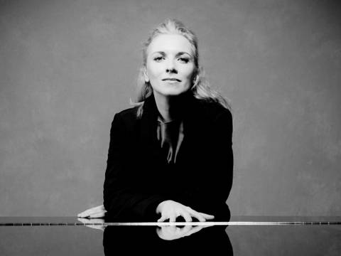 Die Pianistin Tamara Stefanovich – Eine Frau mit blonden Haaren in einem schwarzen Samtblazer steht hinter einem zugeklappten Klavierflügel und schaut in die Kamera.