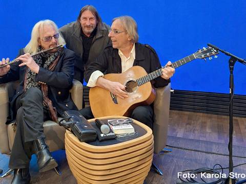 STASI-KNAST & OSTSEEFLUCHT – Drei Personen, zwei sitzen mit Gitarre und Querflöte auf Sesseln, eine steht hinten. Tisch mit Mikrofon, Buch und CD. Blauer Hintergrund, Text: Foto: Karola
