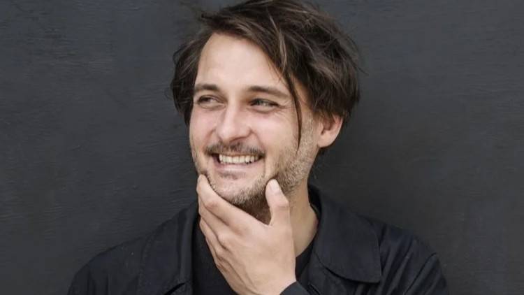 Tilman Rammstedt – Person mit dunklem Haar, schaut nach links, lächelt, hält eine Hand am Kinn, trägt dunkle Jacke vor schwarzem Hintergrund.