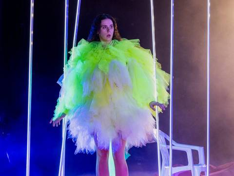 – Person in fluoreszierendem Tüllkleid steht zwischen leuchtenden LED-Streifen; Hintergrund dunkel, seitlich ein weißer Stuhl sichtbar.