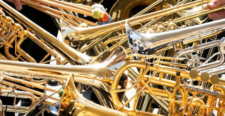 Goldene und silberne Blechblasinstrumente sind dicht ineinander verwoben. Hände mit Trommelstöcken bewegen sich darüber.