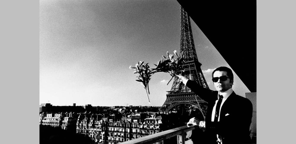 Helmut Newton, Karl Lagerfeld, Paris 1976 – männlich gelesene Person in Anzug hält Lilienstrauss auf Pariser Balkon; Eiffelturm im Hintergrund; Schwarz-Weiß-Fotografie.