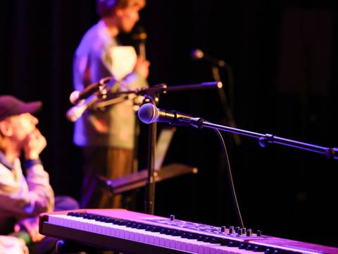  – Im Vordergrund ist ein Mikrofon zu sehen, das über einem E-Piano positioniert ist. Im dunklen Hintergrund sind zwei Personen zu erkennen, von denen eine sitzt und die andere ein Mikrofon in der Hand hält.