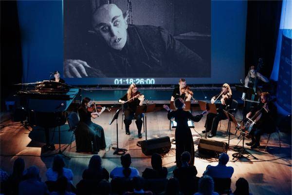 Nosferatu – Orchester spielt vor Leinwand mit projiziertem Vampirfilm. Sechs Musizierende, dirigiert von einer Person, sitzen auf einer Bühne in einem dunklen Raum.