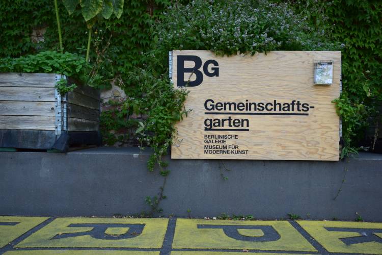 Gemeinschaftsgarten auf dem Gelände der Berlinischen Galerie – Foto: Ein Holz-Schild trägt die Aufschrift „Gemeinschaftsgarten“ und das Logo der Berlinischen Galerie. Um das Schild herum wachsen Pflanzen. Links daneben ist ein weiterer Pflanzkübel zu sehen.