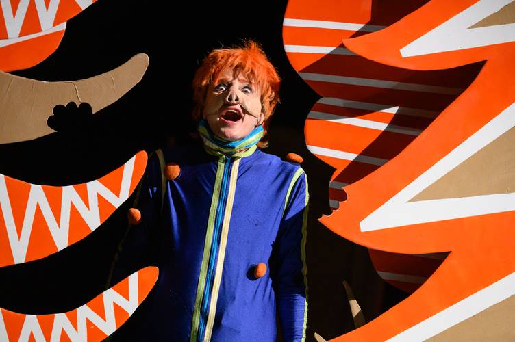Person mit rotem Haar trägt blaues Kostüm mit gelben Streifen. Hintergrund in orange und weiß gemustertem Design.