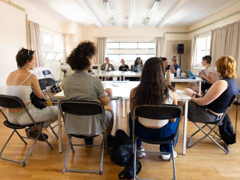 Time to number - Kosten- und Finanzierungspläne – Mehrere Personen sitzen auf Stühlen um einen weißen Tisch in einem Raum mit Holzboden und großen Fenstern.