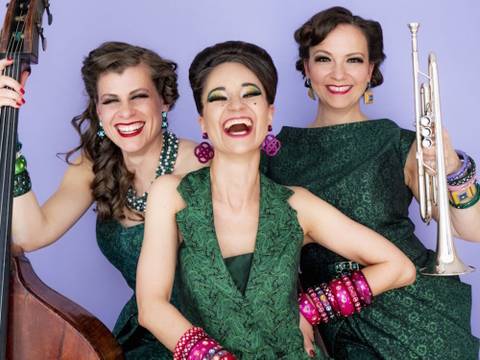Zucchini Sistaz „Eleganz oder gar nicht!“ – Drei lachende Personen, die Instrumente halten: Kontrabass und Trompete. Alle tragen grüne Kleider und farbige Armbänder vor einem lila Hintergrund.