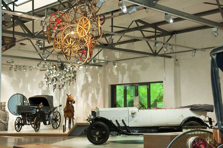 Die Ausstellung zum Straßenverkehr zeigt unter dem Motto „Unterwegs mit Auto & Co“ vielfältige Exponate rund um die Kulturgeschichte der Mobilität. – Blick in die Straßenverkehr-Ausstellung: Ein weißes historisches Cabriolet, eine Kutsche mit Pferd und eine Deckeninstallation aus verschiedenen Holz-Rädern.